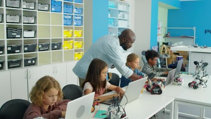 Medium full tracking shot of African American male IT teacher walking around classroom and checking on young diverse students writing programs on laptops for robots. Text in Cyrillic RoboQuantum - Powered by Adobe