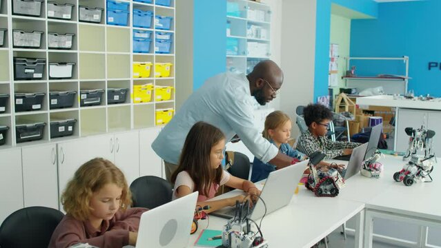 Medium full tracking shot of African American male IT teacher walking around classroom and checking on young diverse students writing programs on laptops for robots. Text in Cyrillic RoboQuantum