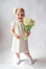Portrait of a smiling little girl in a white dress on white background full-length. Toddler embraces a bouquet of fresh lilies of the valley . Gift for the holiday, the concept of purity, spring time