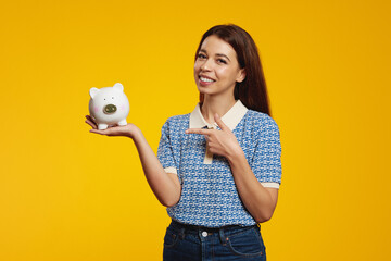 Cute girl in casual blue shirt holding and pointing at white piggy bank with lots of money isolated over yellow background. Money box