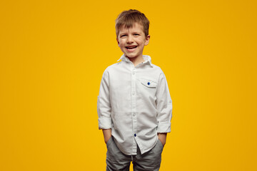 Cute attractive little kid boy in trendy white shirt keeping hands in pockets and smiling for camera against yellow background