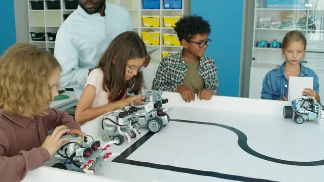 Medium shot of excited nine-year-old diverse children testing their electric robot models at kids hobby club and pressing buttons on control panels, and black male teacher watching and guiding