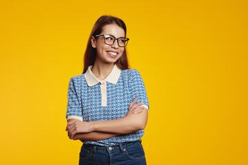 Attractive woman in blue shirt and eyeglasses smiling while looking at free empty space for text with crossed arms, isolated over yellow background