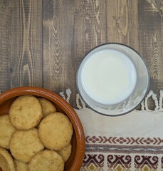 Glass of milk, cookies in traditional clay dish, wooden background and handmade tablecloth, top view.