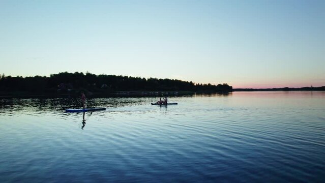 Low angle arc shot flying around woman with kids on sup board on calm lake with water ripples in evening twilight. Silhouettes of the forest and orange purple blue sunset sky