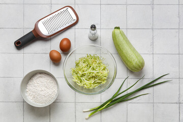 Bowl with grated zucchini and ingredients for preparing fritters on white tile background