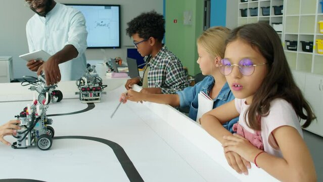 Medium shot of group of young diverse children with African American teacher with tablet watching programmable electric robot models wheel around on table during lesson at after school robotics club