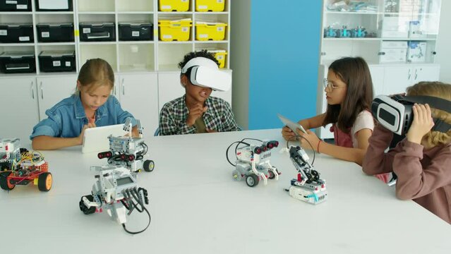 Full shot of young multiethnic children spending time at after school IT club - girls using tablet computers, boys in VR headsets fooling around, and electric robot models on table