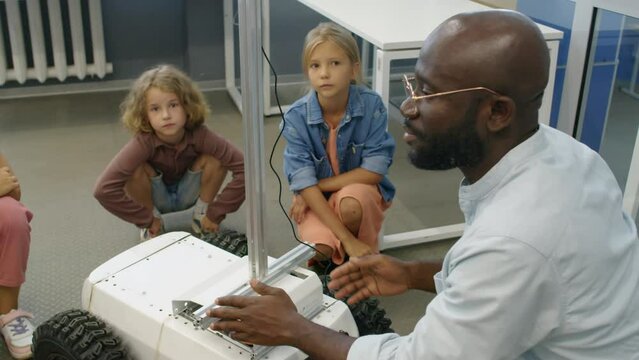 Medium arc shot of young multiethnic boys and girls in casual clothes sitting around delivery robot in classroom, and African American male teacher in glasses explaining its design and functions