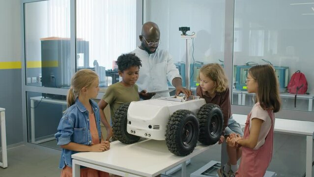 Full shot of group of diverse preteen children standing around delivery robot model in lab, looking at its wires and circuitry, and listening to explanations of African American male teacher