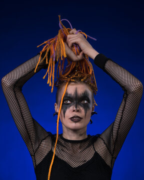 Informal young female with colored braids hairdo and spooky black stage makeup painted on face. Front view, studio shot on blue background. Part of photo series