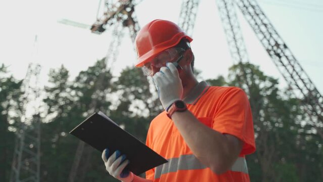 Electrical engineer on high voltage power transformer substation reporting problem with walkie-talkie and looks into clipboard. Professional electrician with portable radio near high voltage tower 