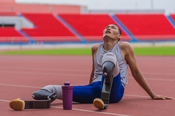 Tired Asian speed runner, equipped with two prosthetic running blades, sits on the stadium track, resting after an intense speed running practice session