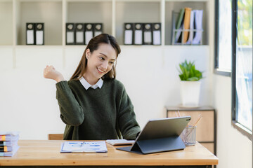 Happy young asian businesswoman celebrating success with arms raised in front of laptop with excitement at office