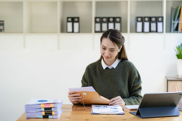 Asian businesswoman in suit sitting on desk in office, with computer document graph for bookkeeping in workplace to calculate annual profit by function, business concept