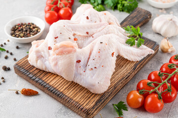 Raw chicken wings on a wooden cutting board with spices, fresh herbs, tomatoes on a gray concrete background. Close up.