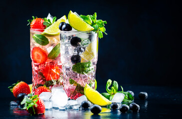 Cold summer strawberry and blueberry mojito cocktail drinks with lime, white rum, soda, cane sugar, mint, and ice in highball glass on deep blue background. Cool refreshing beverages