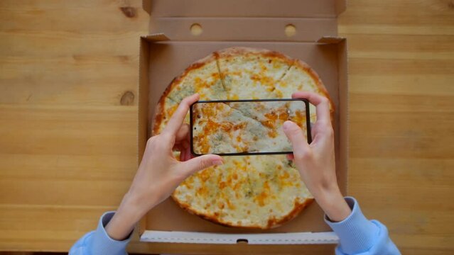 Blogger photographing fresh 4 cheese pizza. Top view - Female hands in blue sweatshirt use smartphone, take photo for social networks appetizing fresh cooked italian Quattro Formaggi Pizza in delivery