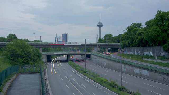 Traffic Flow and Cologne Skyline. Witness bustling cars with light strokes on a highway, a prominent TV tower, moving clouds, and Cologne's panoramic skyline in the evening. Energy and dynamism. 001