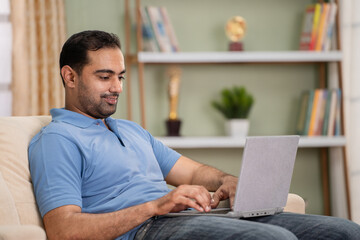 Happy smiling indian man using laptop while sitting on sofa at home - concept of technology, weekend and surfing internet.