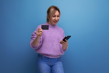 portrait of a beautiful blonde young woman in a lilac sweater making an online purchase holding a credit card with a mockup on a bright background with copy space
