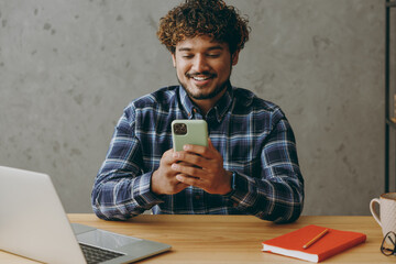 Successful smiling happy fun cheerful young employee business Indian man he wears casual blue checkered shirt hold in hand use mobile cell phone sit work at office desk with laptop pc computer indoor