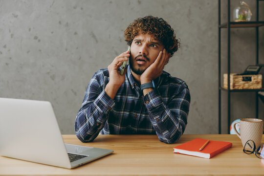 Preoccupied sad upset worried young employee business Indian man wearing casual blue checkered shirt talking speak on mobile cell phone look asit work at office desk with laptop pc computer indoors.