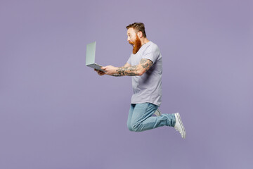 Full body young redhead bearded IT man wear violet t-shirt casual clothes jump high hold use work...