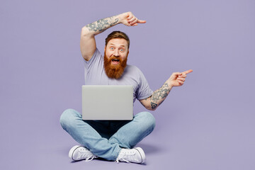 Full body young redhead bearded man in violet t-shirt casual clothes sitting hold use work on...