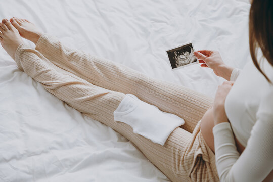 Cropped sideways young pregnant woman in white pyjamas hold little clothes picture of ultrasound test rest relax spend time in bedroom room lounge home Maternity family pregnancy expectation concept