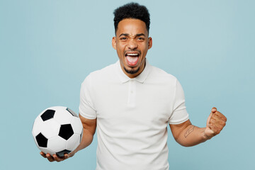 Young man fan wear basic t-shirt cheer up support football sport team hold in hand soccer ball watch tv live stream do winner gesture clenching fists isolated on plain pastel blue background studio.