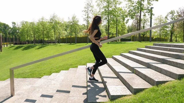 A young beautiful woman with long hair in black sportswear runs up the steps in a modern park. The camera moves slowly along the runner with the stabilizer.