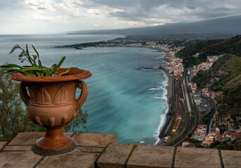 Mediterranean flowering plant in a stone ceramic pot with scenic view on seaside and Mount Etna volcano from public garden Parco Duca di Cesaro to Giardini Naxos in Taormina, Sicily, Italy