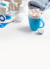 The blue cup with warm chocolate and marshmallows on the top; clew, pompom, and gift boxes, scotch tape, ribbons, and scissors. There is space for text.