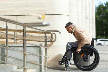 Side view of young man sitting in wheelchair using ramp to move up of building