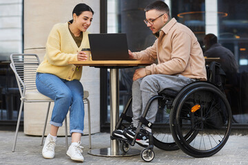 Young man with disability having business meeting with woman, they sitting in outdoor cafe and discussing online presentation on laptop