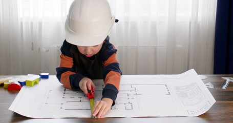 Preschooler in hardhat measures dimensions with tape measure on apartment drawing. Boy imitates role of engineer taking measurements with interest using tool on table