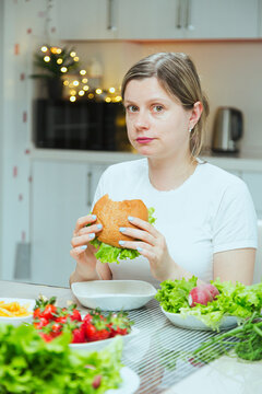 Young woman eating hamburger in the kitchen with full table of different food, problems with bulimia. Breaking down diet and fitness, say no to stereotypes and diets.