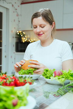 Young woman eating hamburger in the kitchen with full table of different food, problems with bulimia. Breaking down diet and fitness, say no to stereotypes and diets.