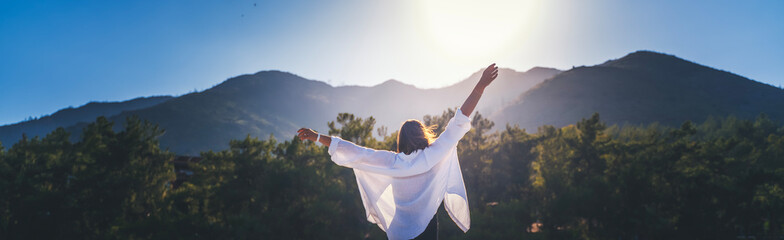 A woman in a white shirt with her hands raised up enjoy the morning sun against the backdrop of...