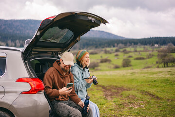 Romantic happy middle age caucasian couple traveling by car sitting in open trunk with photo camera and smartphone - 615712397