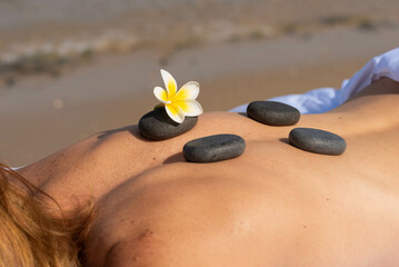 Obraz na płótnie Canvas Massage hot stones on woman’s back with tropical flower. Woman having relax in tropical massage spa on the beach near the sea on massage table.