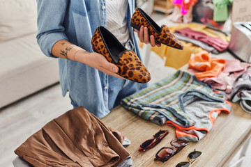 ethical consumption, exchange, cropped view of young tattooed woman holding stylish animal-print shoes near sunglasses and garments on table, sustainable living and mindful consumerism concept