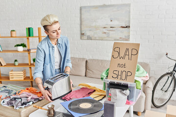 young tattooed woman with electric toaster near vinyl record player, second-hand clothes and carton card with swap not shop lettering at home, sustainable living and circular economy concept