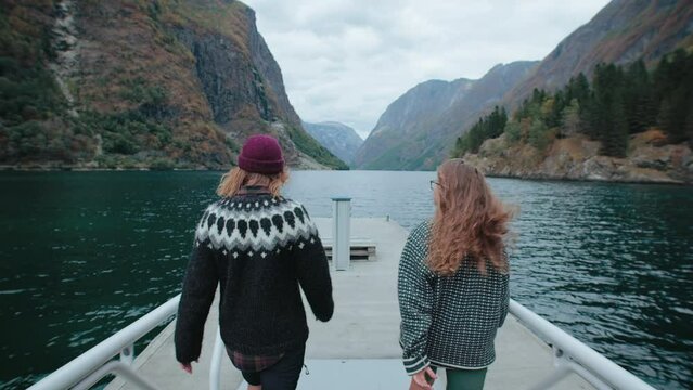 Two female friends in authentic traditional scnadinavian sweaters walk on pier or boardwalk near fjord or glacier lake. Look in awe on amazing nordic landscape