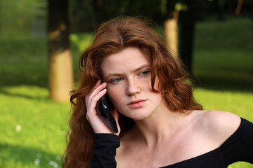 Attractive girl with long red hair and freckles talking on mobile phone sitting on a bench in summer park