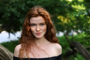 Naklejka premium Portrait of pretty ginger girl with long red hair and freckles standing in summer park. Young sensual model, natural female beauty