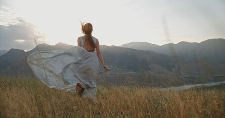 Fototapeta na wymiar Young beautiful girl with red hair wearing white dress running on top of a mountain facing wind blowing her hair and dress and smiling - freedom, adventure, harmony 
