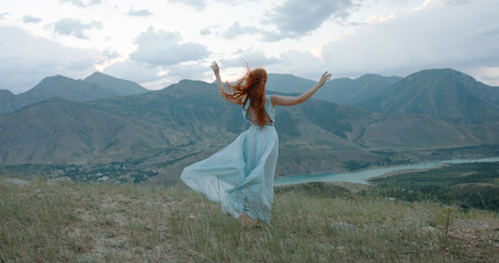Fototapeta na wymiar Beautiful carefree girl with red hair wearing white dress standing against the wind, looking at mountains and raising her hands - freedom, adventure, inspiration 