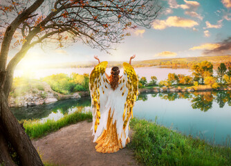 Art photo Fantasy woman goddess angel raises hands to blue sky golden white wings long gold dress standing on top of mountain pray back rear view on river magic divine sun light. Fairy girl sexy lady